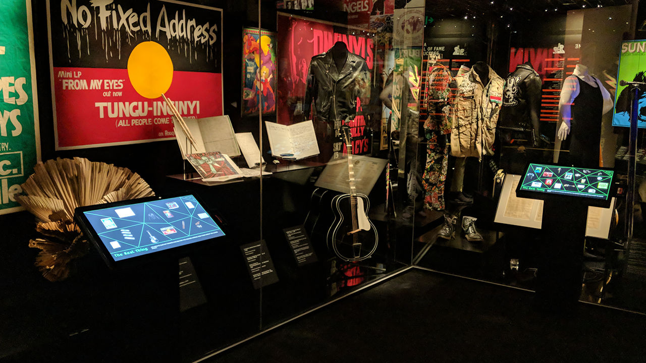 Wideshot of exhibition containing guitars, outfits and flags behind glass case.