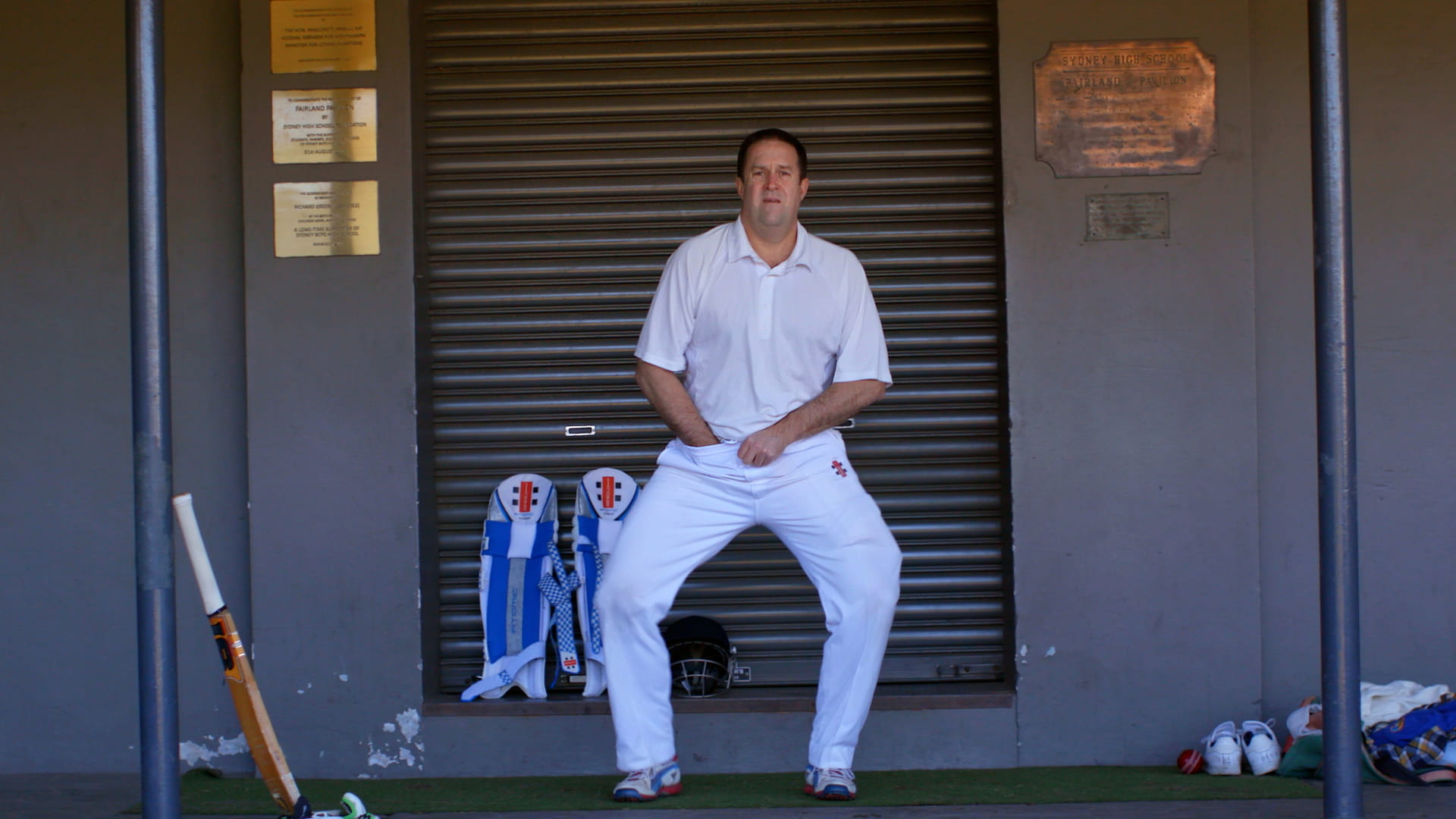 Male cricketer adjusting his box next to a Sydney Highschool plaque.